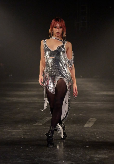 Sonbahar/Kış 2025 Kopenhag Moda Haftası defilelerinde Alectra Rothschild'ın maskülen ve feminen ögeleri harmanlayan Masculina koleksiyonunu sergileyen, kırmızımsı-sarı saçlı bir model, parçalı ve pırıltılı gümüş payetli elbisesi, siyah külotlu çorabı ve zımbalı botlarıyla podyumda iddialı bir duruş sergiliyor.