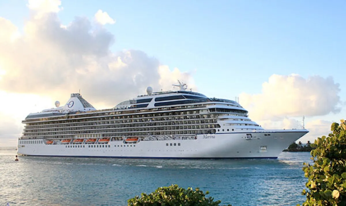 Mavi denizde süzülen, beyaz gövdeli ve çok katlı Oceania Cruises Marina yolcu gemisi, yeşil bitki örtüsüne sahip kıyı şeridinin ve hafif bulutlu gökyüzünün önünde görünüyor, lüks bir cruise deneyimini yansıtıyor.