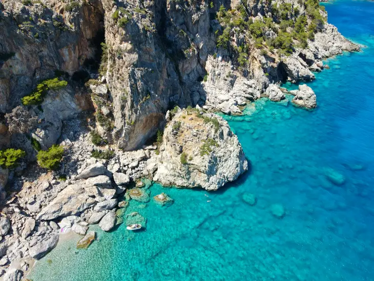 Olympos'un masmavi sularla buluştuğu sarp kayalık kıyı şeridinin havadan çekilmiş bu nefes kesici görüntüsünde, berrak denizde demirlemiş küçük bir tekne ve yüzen birkaç kişi, yemyeşil bitki örtüsüyle kaplı dik yamaçların doğal güzelliğini vurguluyor.