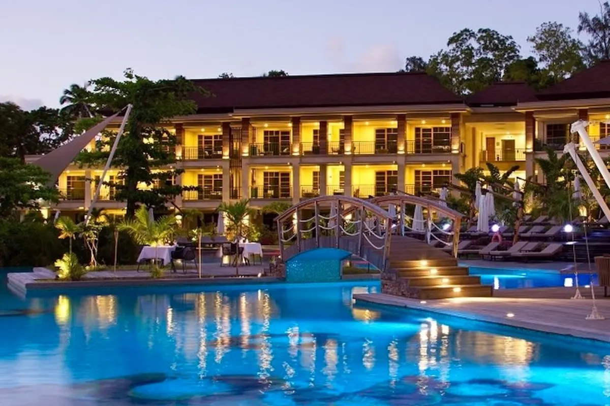 Savoy Seychelles'in etkileyici lüks tatil köyü manzarasında, gece ışıklarıyla parlayan çok katlı bina, aydınlık pencereleri ve balkonlarıyla dikkat çekerken, ön plandaki turkuaz rengi yüzme havuzu, ışıkları yansıtan yüzeyi ve üzerinde yer alan şık ahşap köprüyle tropik bitkilerin arasında yer alan dış mekan yemek alanlarına eşlik ediyor; en güzel balayı rotaları için ilham veren bir kare.