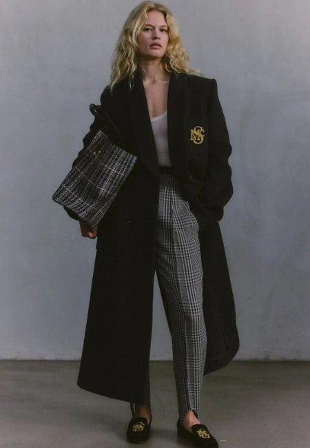 Siyah oversize kaban, kareli pantolon ve monogram detaylı loafer ayakkabılarla tamamlanan modern kadın kış kombini, güçlü ve sofistike şehir stili görünümü.