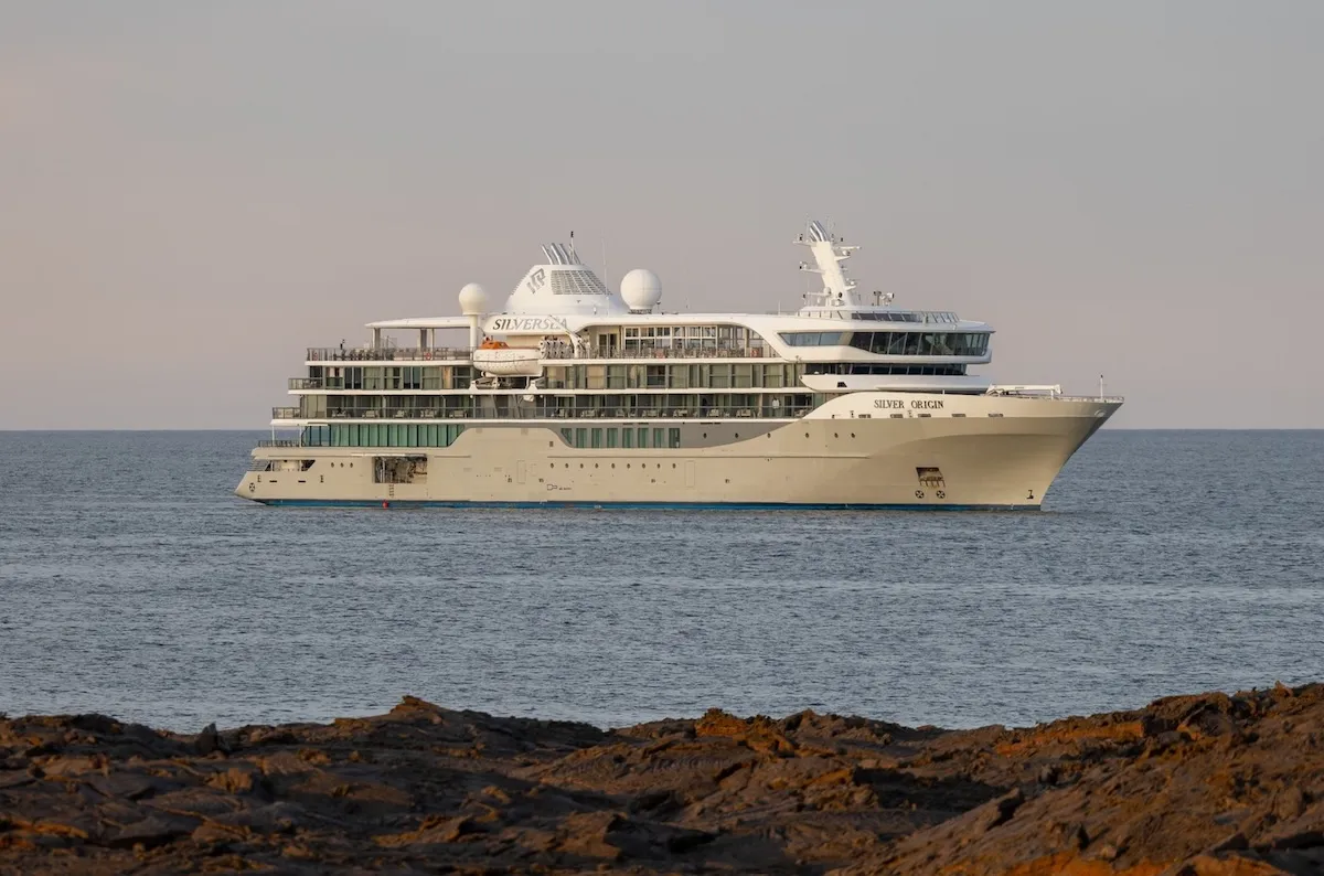 Uzakta koyu renkli kayalık bir kıyı şeridinin önünde, sakin mavi denizde lüks Silversea kruvaziyer gemisi SILVER ORIGIN.