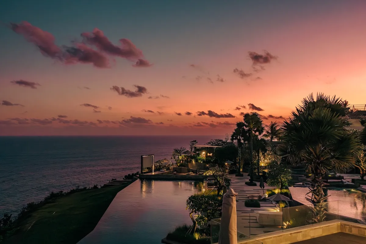 Gün batımının okyanus üzerinde pembeye, turuncuya ve mora çalan gökyüzüyle birleştiği, Bali'deki Six Senses Uluwatu'nun lüks tatil köyündeki aydınlatmalı sonsuzluk havuzunun ve palmiye ağaçlarının eşlik ettiği büyüleyici bir akşam manzarası.