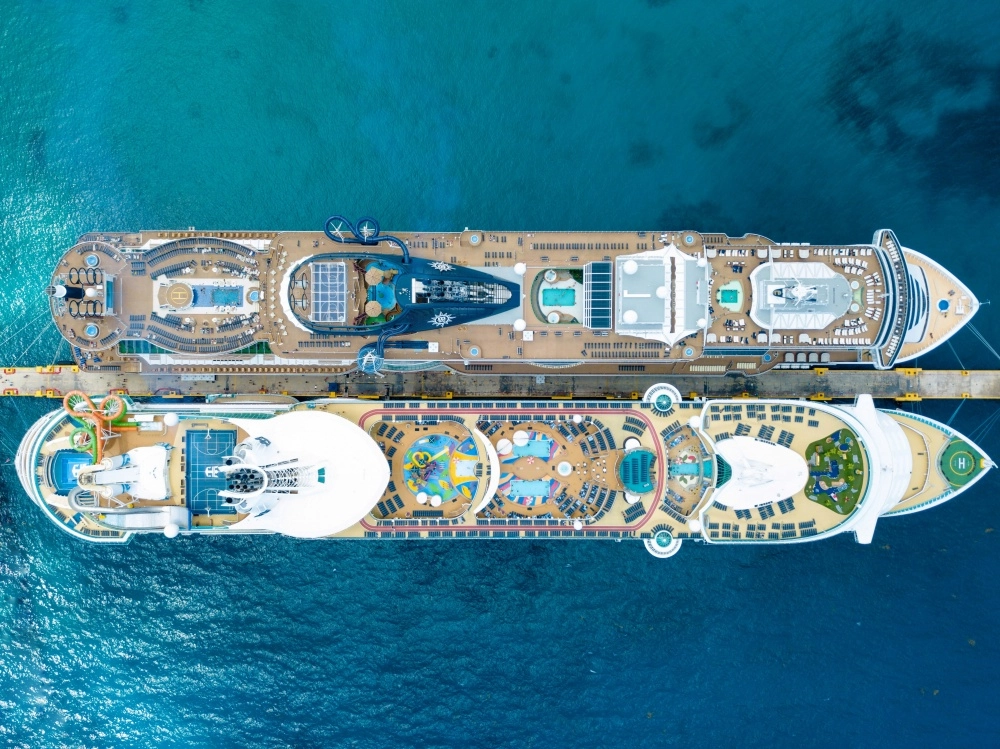 Drone çekimiyle çekilmiş bu kuşbakışı fotoğrafta, turkuaz renkteki berrak sularla çevrili bir iskeleye yan yana yanaşmış iki devasa ultra-lüks cruise gemisi görülüyor; gemilerin üst güvertelerinde havuzlar, çeşitli su kaydırakları, spor sahaları ve yemyeşil dinlenme alanları gibi birçok eğlence ve dinlence imkanı detaylı bir şekilde sergileniyor.