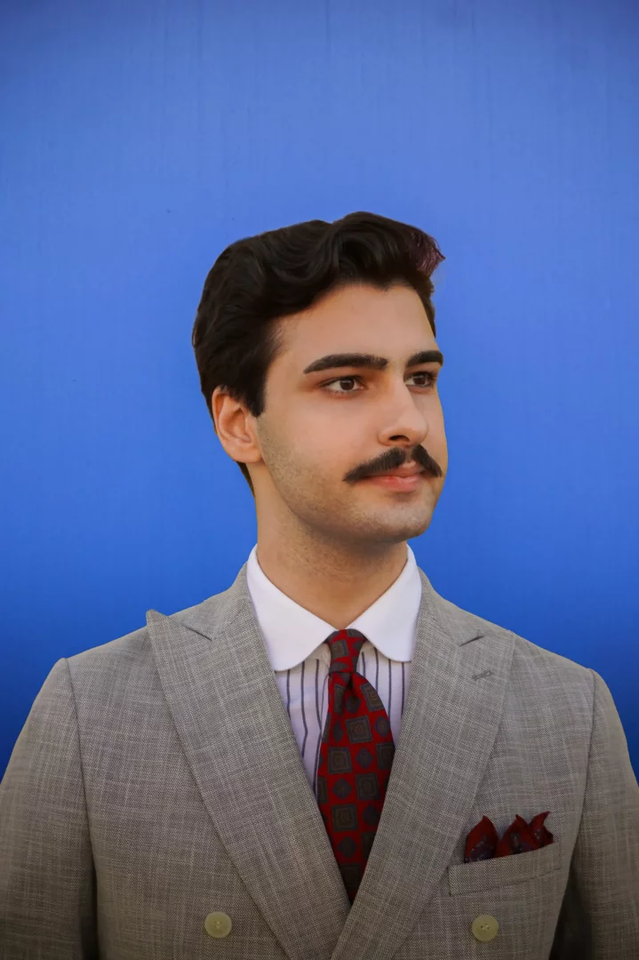Umut Kaya'nın lacivert arka plan önünde, dalgalı koyu saçları, özenle şekillendirilmiş bıyığı, gri kruvaze ceket, çizgili gömlek, desenli kırmızı kravat ve cep mendiliyle, zarif bir şekilde sağa doğru baktığı profesyonel bir portre fotoğrafı.