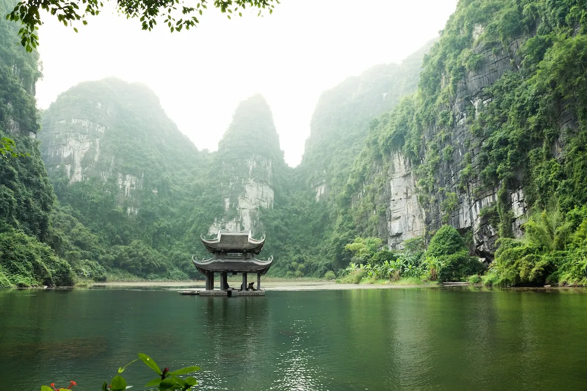 Vietnam'ın yemyeşil karst dağlarıyla çevrili, sisli bir havada parlayan sakin bir su kütlesinin ortasında, zarif mimariye sahip ahşap bir pagoda yapısı yükseliyor, doğanın ve kültürün buluştuğu huzurlu bir manzara sunuyor; en güzel balayı rotaları için ilham veren bir kare.