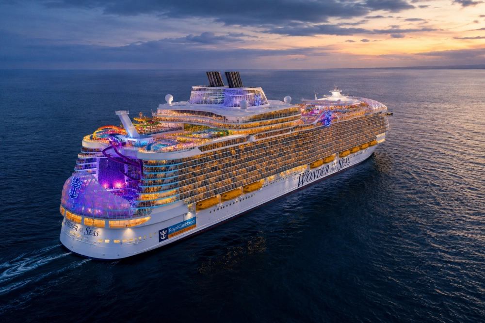 Wonder of the Seas adlı Royal Caribbean cruise gemisi, mor ve turuncu ışıklarla aydınlanmış güverteleri ve su kaydıraklarıyla, gün batımının kızıl ve mavi gökyüzü altında denizde süzülerek ultra lüks bir cruise tatili deneyimi sunuyor.
