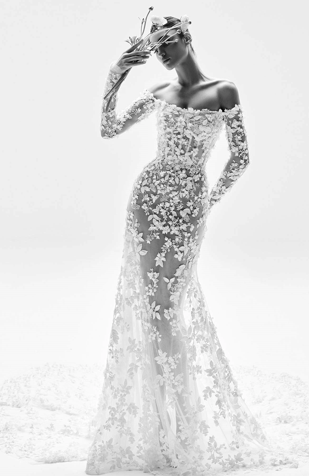 Zuhair Murad'ın 2025 İlkbahar gelinlik koleksiyonundan, şeffaf kumaş üzerine zarif üç boyutlu çiçek işlemeleriyle bezenmiş, omuzları açık ve vücudu saran ince belli bir gelinlik giymiş model, elinde çiçekli bir aksesuar tutuyor.