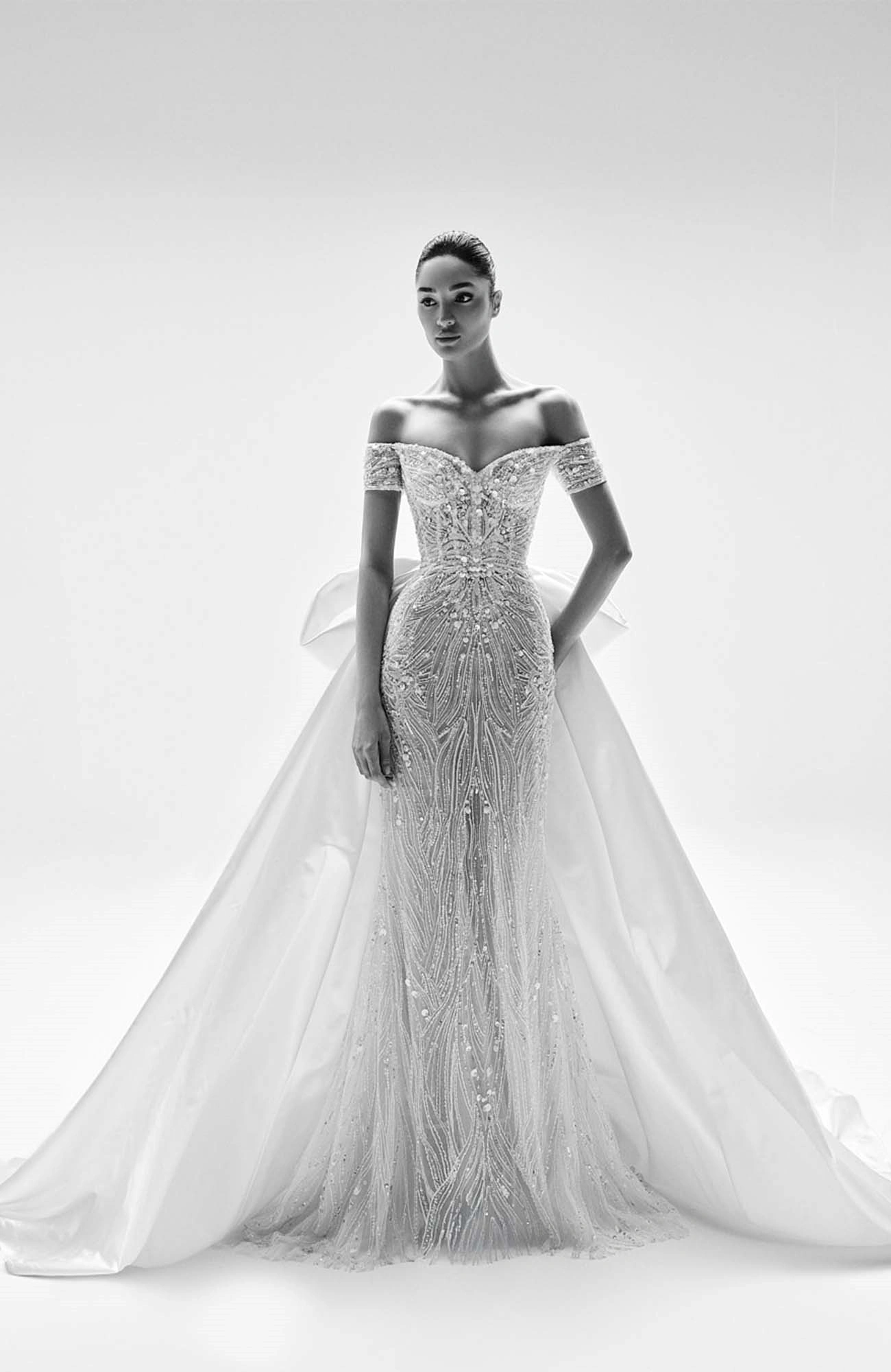 Zuhair Murad 2025 İlkbahar gelinlik koleksiyonundan, omuzları açık, vücuda oturan ve Fenike mirasına gönderme yapan bitki motifli, ışıltılı işlemelerle bezeli bu zarif model, arkasındaki geniş, akışkan kuyruk detayıyla antik bir efsaneden fırlamışçasına bir zarafet sunuyor.