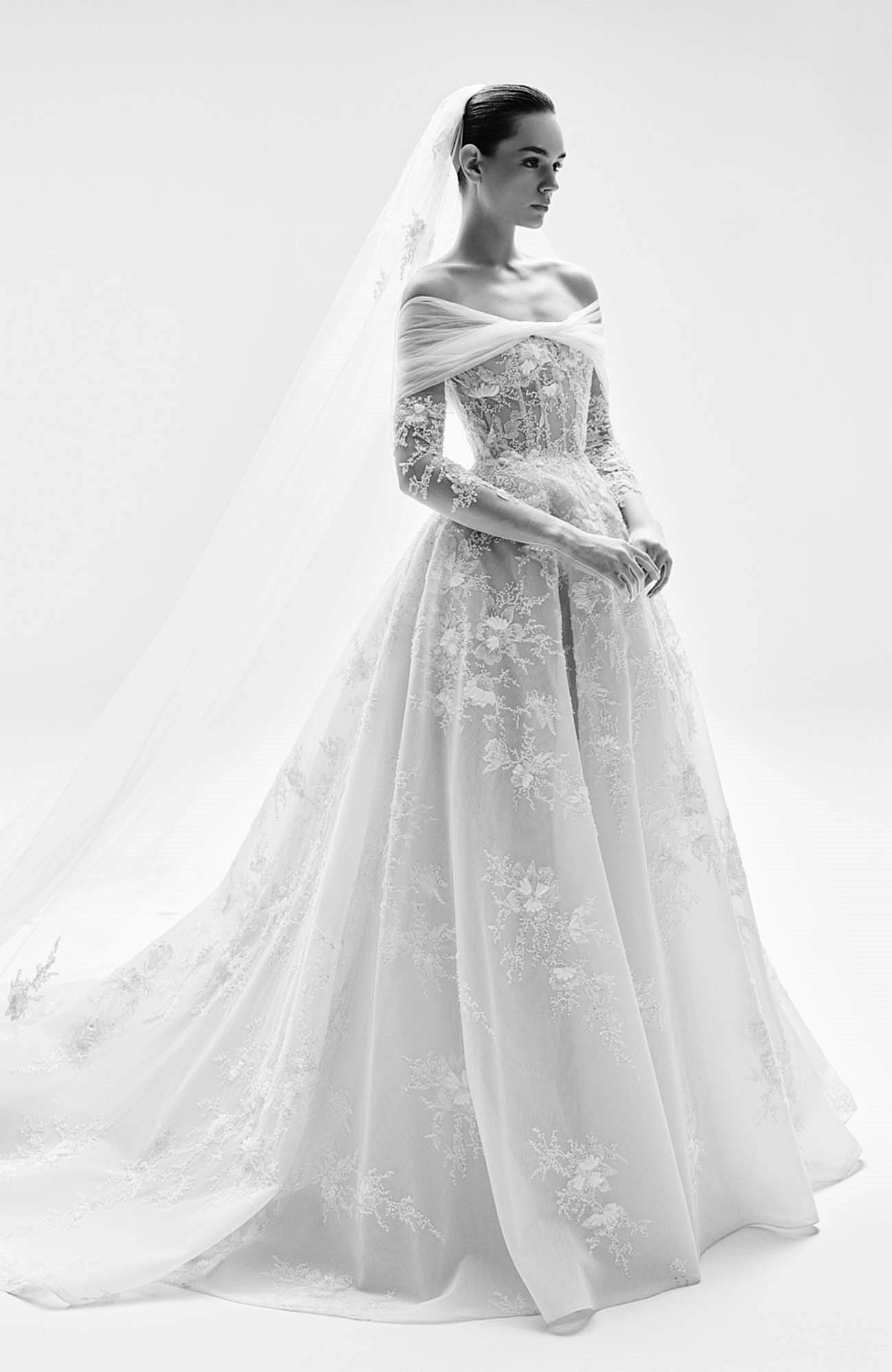 Zuhair Murad 2025 İlkbahar gelinlik koleksiyonundan, omuzları açık, uzun kollu, transparan dokular üzerinde zarif üç boyutlu çiçek işlemeleri ve dantellerle bezenmiş, hacimli etekli ve uzun duvaklı bir gelinliği zarafetle sergileyen model, markanın couture işçiliğini gözler önüne seriyor.