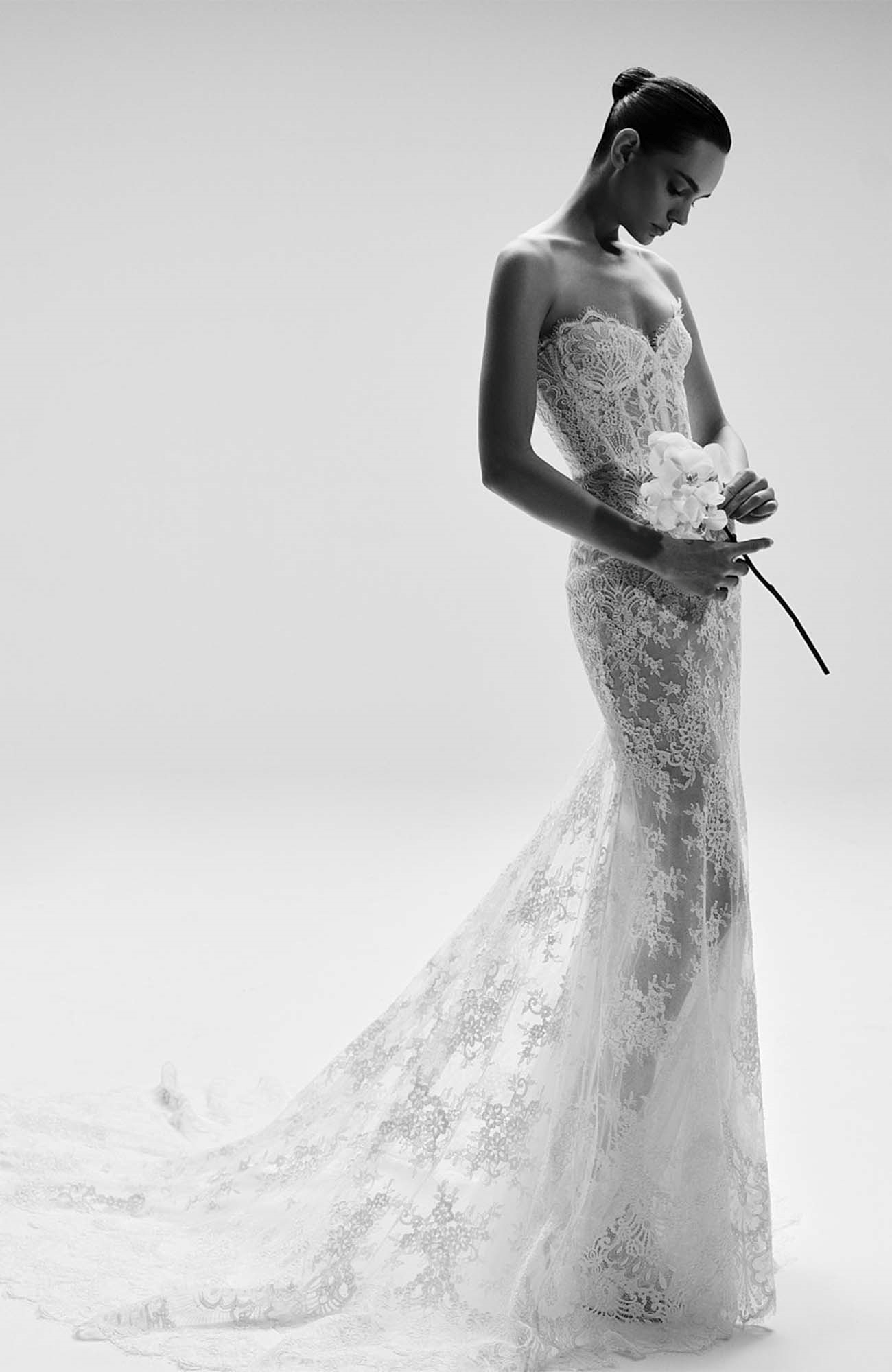 Zuhair Murad 2025 İlkbahar Gelinlik Modelleri koleksiyonundan, zarif dantel işlemeli, bedeni saran straplez beyaz bir gelinlik giyen model, yere değen uzun etek ucuyla romantik bir duruş sergiliyor ve elinde beyaz orkideler tutuyor.