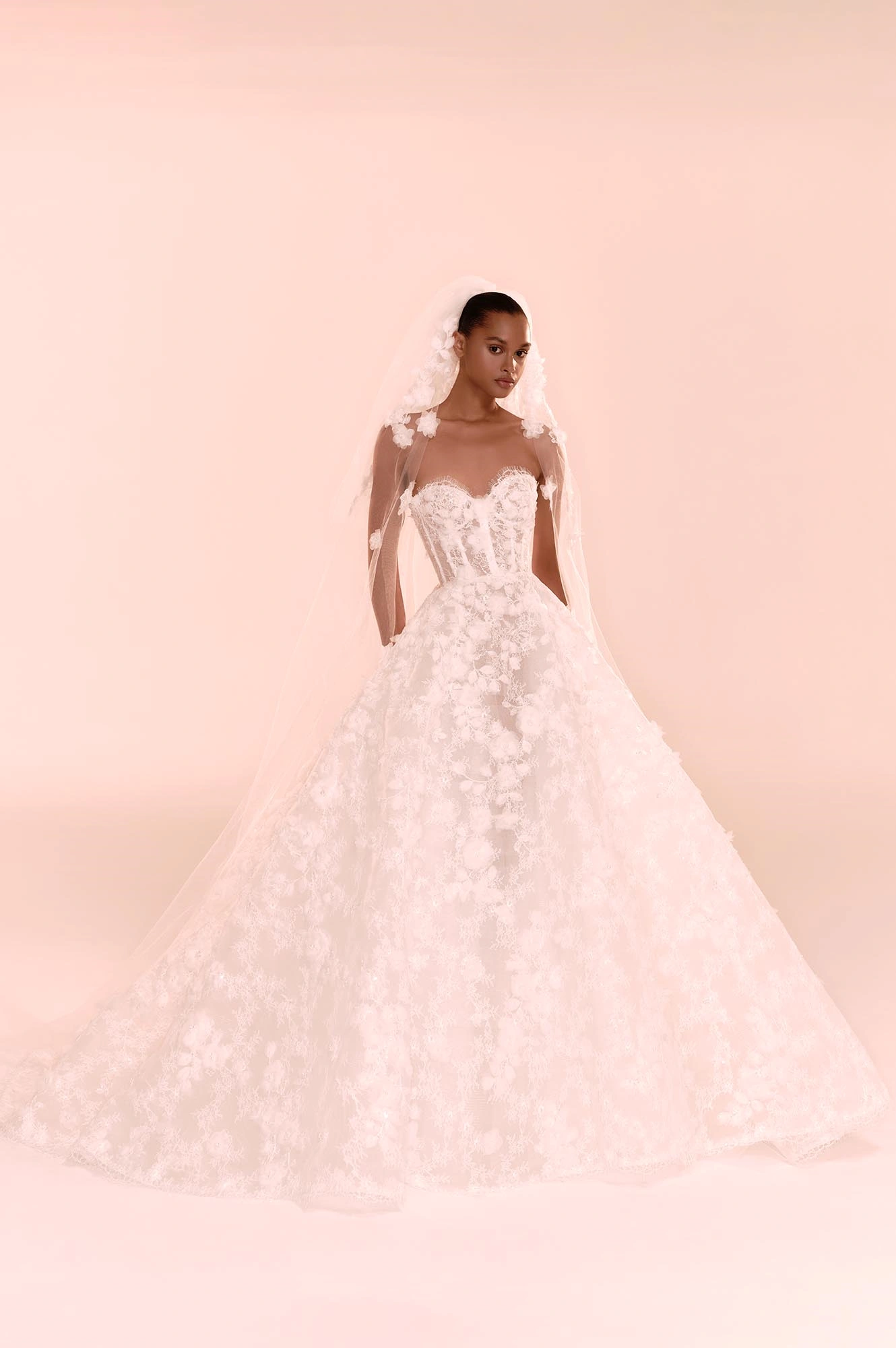 Zuhair Murad'ın 2025 Sonbahar Gelinlik Koleksiyonu'ndan, dantel işlemelerle ve üç boyutlu çiçek aplikeleriyle bezenmiş, korse detaylı kalp yakalı ve kabarık etekli romantik bir gelinlik giyen model, zarif çiçekli tülüyle adeta masalsı bir havayı yansıtıyor.