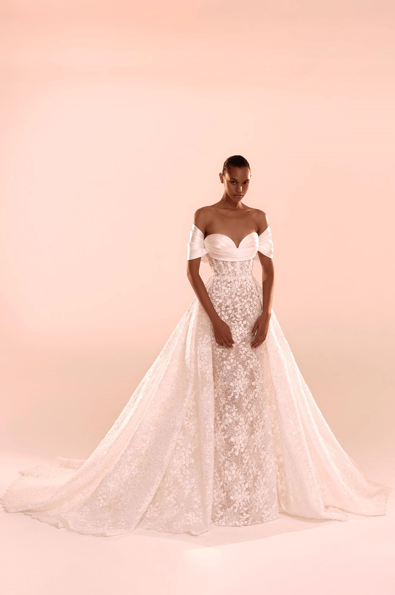 Zuhair Murad'ın 2025 Sonbahar Gelinlik Koleksiyonu'ndan, omuzları nazikçe açıkta bırakan drapeli saten kalp yakası, zarif çiçek aplikeleriyle işlenmiş vücuda oturan iç elbisesi ve ışıkla hareket eden kristallerle bezeli, hacimli ayrılabilir dış eteğiyle romantik ve modern bir gelinlik sergileyen model.