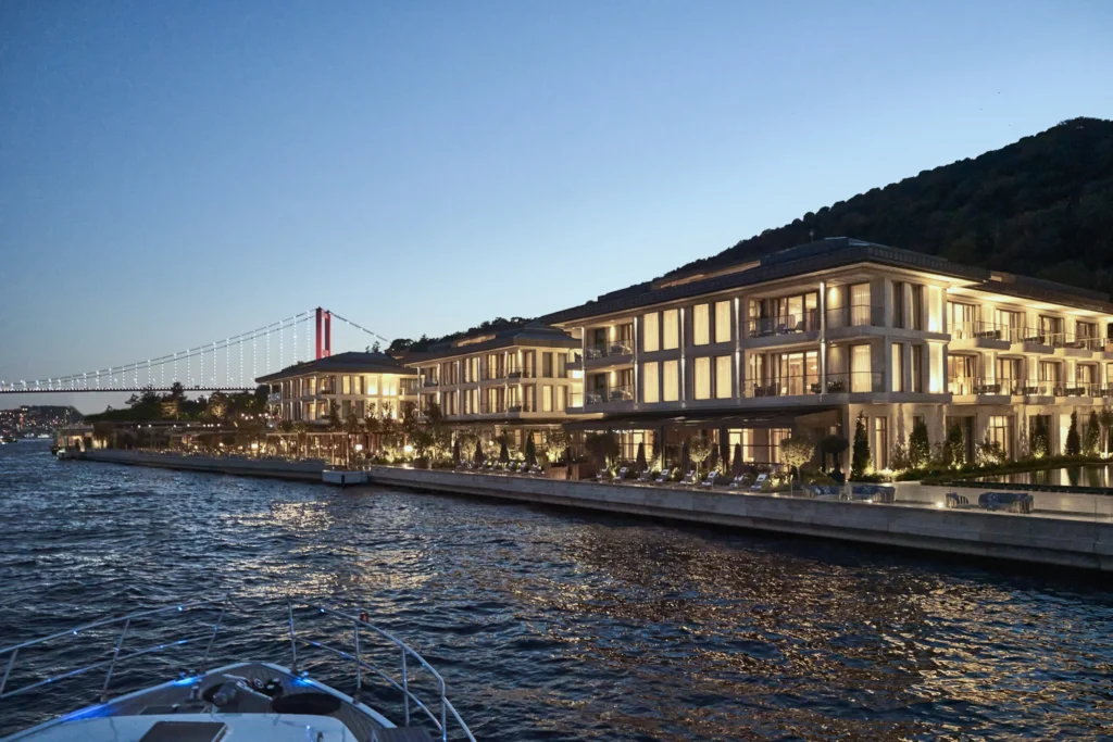 Akşam karanlığında Boğaz'ın ışıklı suları üzerinde yükselen Mandarin Oriental Bosphorus Istanbul oteli, modern mimarisi ve pencerelerinden sızan sıcak ışıklarıyla göz kamaştırırken, geride ışıltılı Boğaz Köprüsü'nün silüeti bir tekneden büyüleyici bir İstanbul manzarasını tamamlıyor.