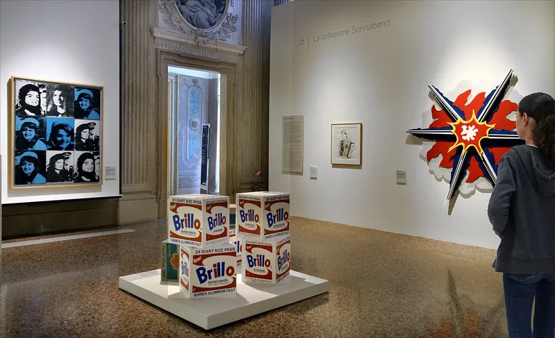 Andy Warhol’un Brillo sabun kutuları yerleştirmesi ve pop art portreleriyle çevrili bir müze salonu; tüketim estetiğini yücelten sergi düzeni.