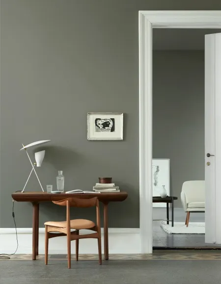 Modern minimalist bir ofis köşesinde, zeytin yeşili duvar ve ceviz ağacı masa üzerinde yer alan beyaz masa lambasıyla sade ve odaklanmayı teşvik eden bir dekorasyon stili.