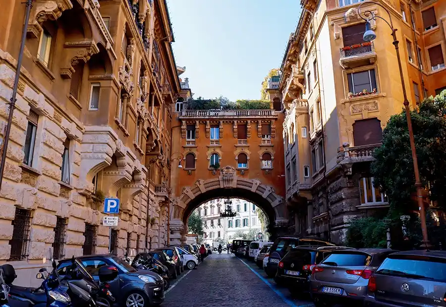 Roma Seyahat Rehberi kapsamında, İtalya’nın Gaudi’si olarak bilinen mimar Gino Coppedé tarafından tasarlanmış, Art Nouveau ve Art Deco tarzlarının Yunan, Gotik ve Barok detaylarla harmanlandığı, havadan asılı avizelerin görüldüğü kemerli bir sokakta sıralanmış süslü binalar arasında park etmiş araçlar ve motosikletler.