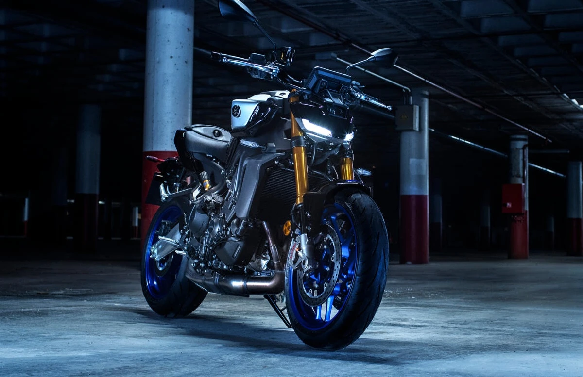 Karanlık bir otoparkta, ön farları açık ve dinamik tasarımıyla dikkat çeken, parlak mavi jantları ve altın rengi amortisörleriyle güçlü bir duruş sergileyen Yamaha MT-09 SP naked motosiklet, beton sütunlar arasında park edilmiş haliyle görünüyor.