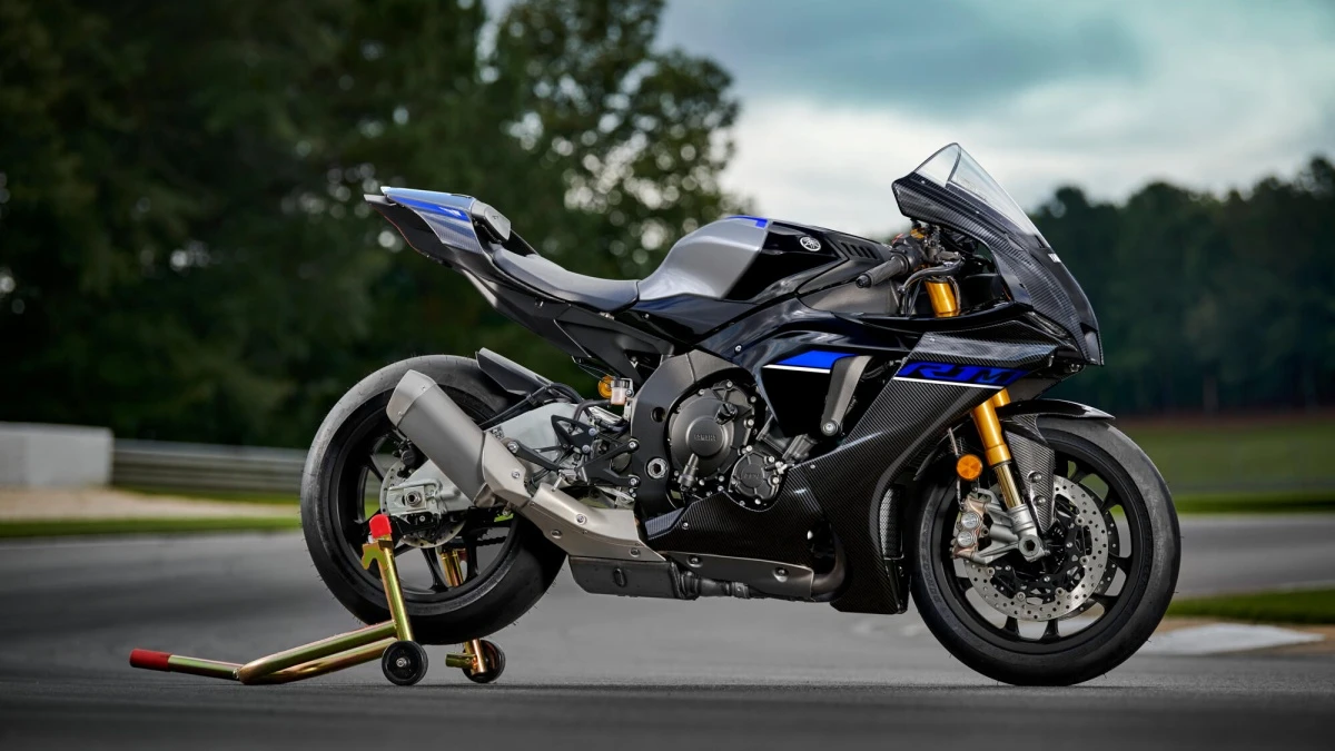 Yamaha YZF1000 R1M yarış motosikleti, mat siyah ve parlak mavi detaylarla bezeli, karbon fiber kaplamalı grenajlarıyla asfalt bir pist yüzeyinde sarı renkli bir sehpada durarak, hıza hazır güçlü bir duruş sergileyen yüksek performanslı bir modeldir.