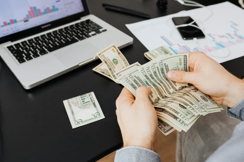 Bir kişi, masa üstünde laptop ve grafikler varken elindeki Amerikan dolarlarını sayıyor.
