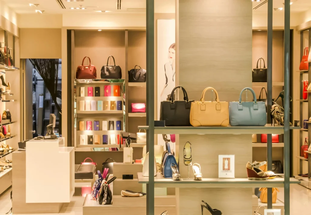 Lüks bir butik mağazada sergilenen çeşitli çanta, ayakkabı ve cüzdan modelleri; raflarda düzenli şekilde yerleştirilmiş kadın aksesuarlarıyla modern bir alışveriş deneyimi sunuluyor.