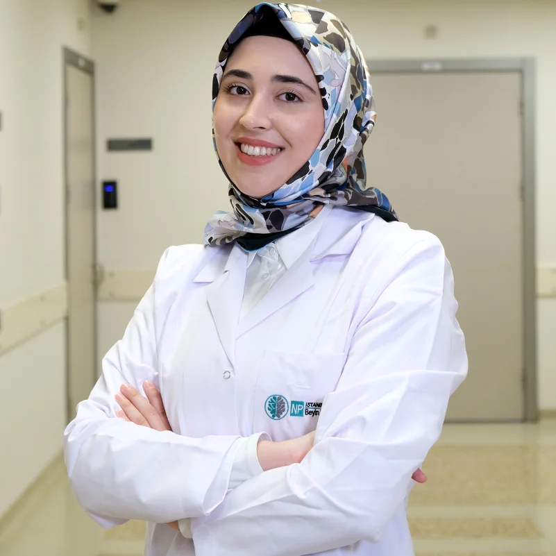 Uzman Klinik Psikolog Zeynep Betül Alp, beyaz önlüğü ve desenli başörtüsüyle hastane koridorunda gülümseyerek kollarını kavuşturmuş, kaliteli uyku için bilinçaltının ve rüyaların önemini vurgulayan bilim destekli ipuçları sunuyor.