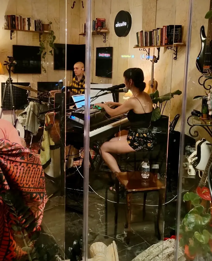Bir stüdyo ortamında sahne alan Ahzuita grubundan iki müzisyen: ön planda piyano çalan ve mikrofon önünde oturan kadın ile arkada elektronik müzik setupı başında oturan adam, Ankara performanslarından birinde müziklerini icra ediyorlar.