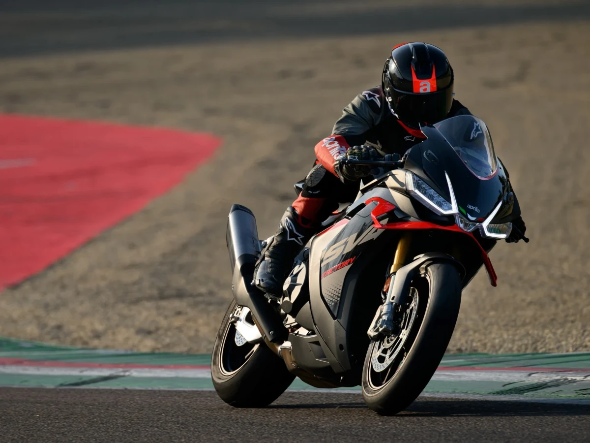 Siyah, beyaz ve kırmızı renk detaylarına sahip Aprilia RSV4 Factory yarış motosikleti, siyah kasklı ve kırmızı detaylı yarış tulumu giyen bir sürücü tarafından, arka planda kırmızı beyaz şeritli virajlı bir yarış pistinde yatık bir şekilde sürülüyor.