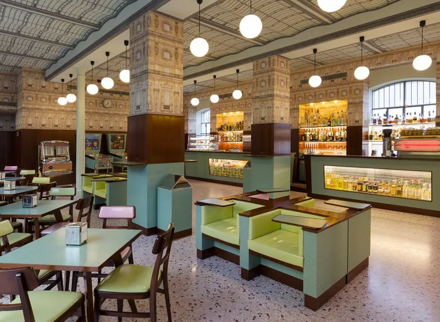 Milano'daki Fondazione Prada bünyesindeki Bar Luce, ünlü yönetmen Wes Anderson'ın yaratıcı dokunuşlarıyla tasarlanmış, mint yeşili masaları, sandalyeleri, koltukları ve desenli sütunlarıyla retro bir atmosfer sunan şık bir müze restoranı; solda bir langırt makinesi ve müzik kutusu, arka planda ise şişelerle dolu bir bar alanı görülüyor.