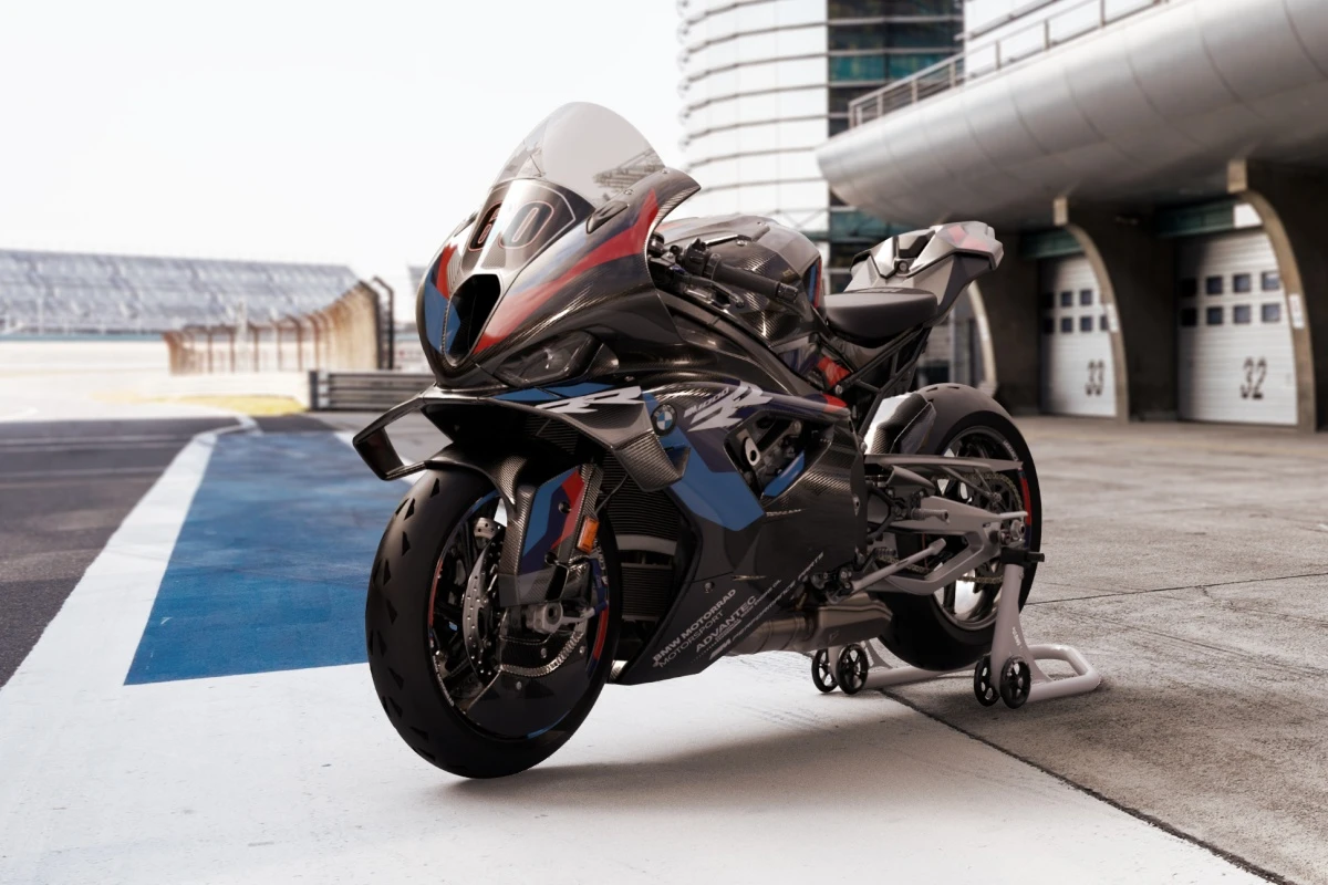 Pist kenarında özel bir sehpa üzerinde sergilenen, aerodinamik karbon fiber gövdesi ve mavi-kırmızı şerit detaylarıyla dikkat çeken, yüksek performanslı, parlak siyah bir BMW M1000 RR yarış motosikleti.