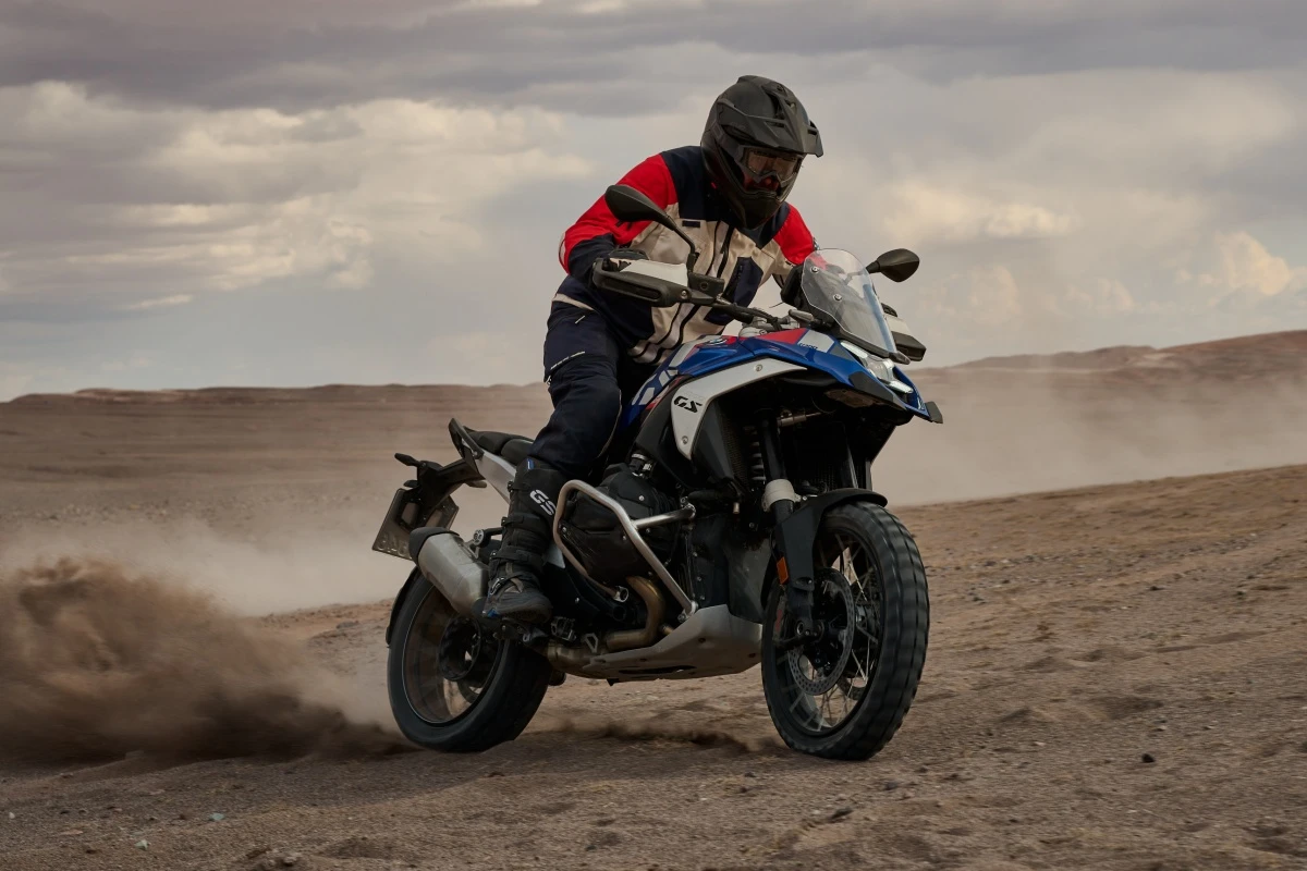 Kask ve tam koruyucu ekipman giymiş bir sürücünün, kumlu, engebeli bir offroad arazide tozu dumana katarak ilerlediği, kırmızı, beyaz ve mavi renk detaylarına sahip BMW R 1300 GS gezi motosikleti tüm ihtişamıyla görünüyor.