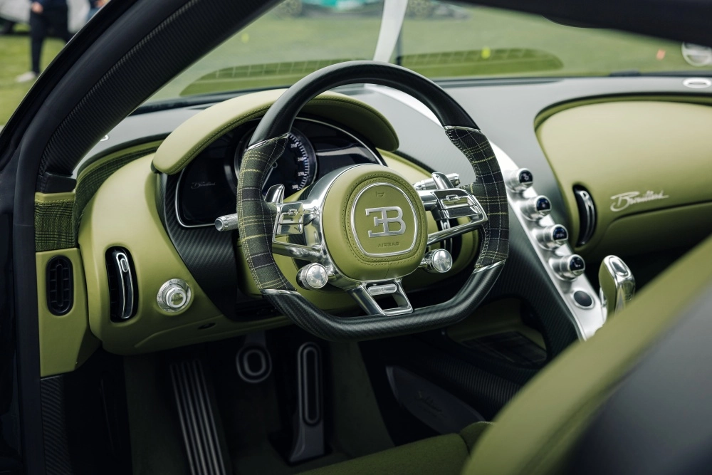 Bu Bugatti Brouillard hiper otomobilin benzersiz yeşil iç mekan tasarımını vurgulayan yakın çekim görselde, sportif detaylara sahip ekose desenli direksiyonun ortasında Bugatti logosu ve lüks gösterge paneli belirgin şekilde görünüyor.