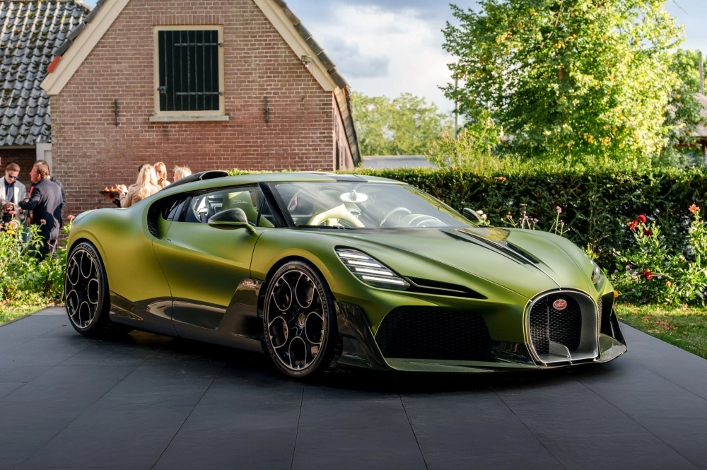 Matte zeytin yeşili renkteki Bugatti Brouillard Programme Solitaire hiper otomobil, tuğla bir binanın ve insanların bulunduğu bir etkinliğin önündeki koyu renkli zeminde park edilmiş, 20 milyon dolarlık bu aracın göz alıcı ön tasarımı ve siyah jantları belirgin bir şekilde görülüyor.