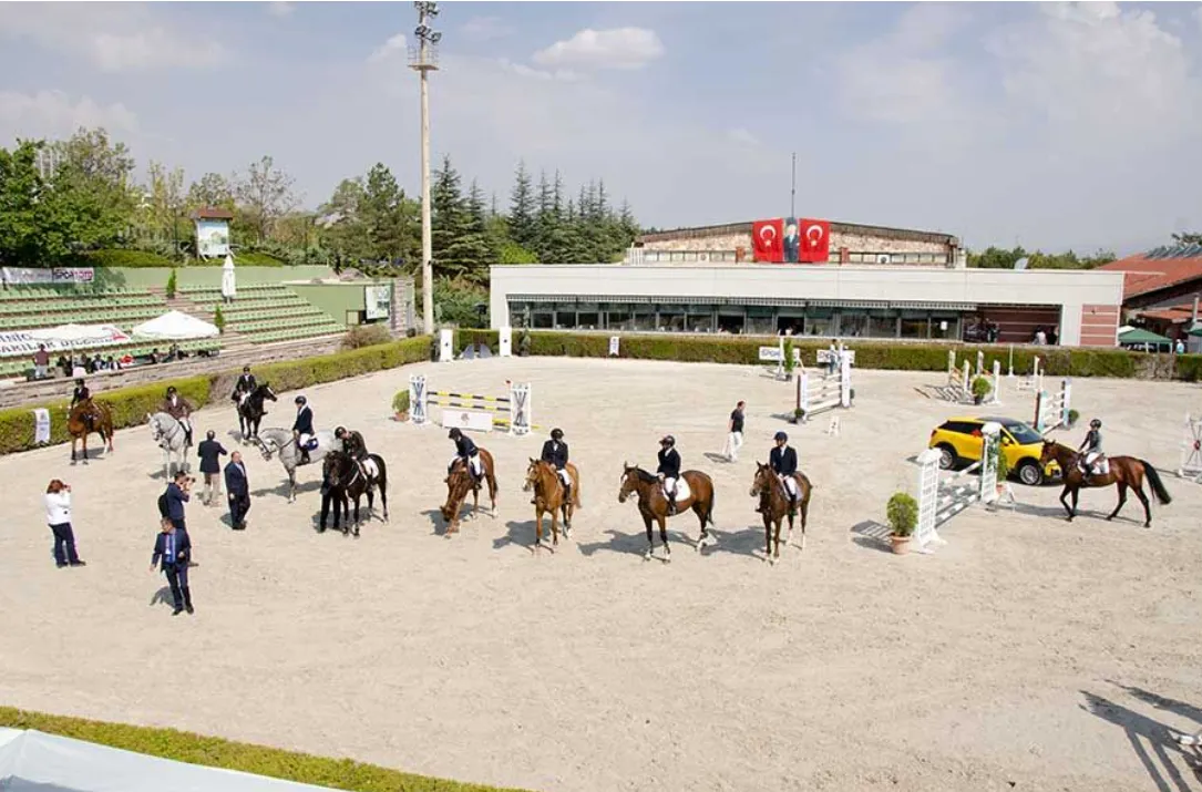 Güneşli bir havada, kumlu arenada birçok atlı ve atın, yeşil tribünler ve Türk bayraklarının asılı olduğu kulüp binası önünde at biniciliği etkinliği veya engel atlama yarışması için toplandığı geniş bir açık hava tesisi görülmektedir.