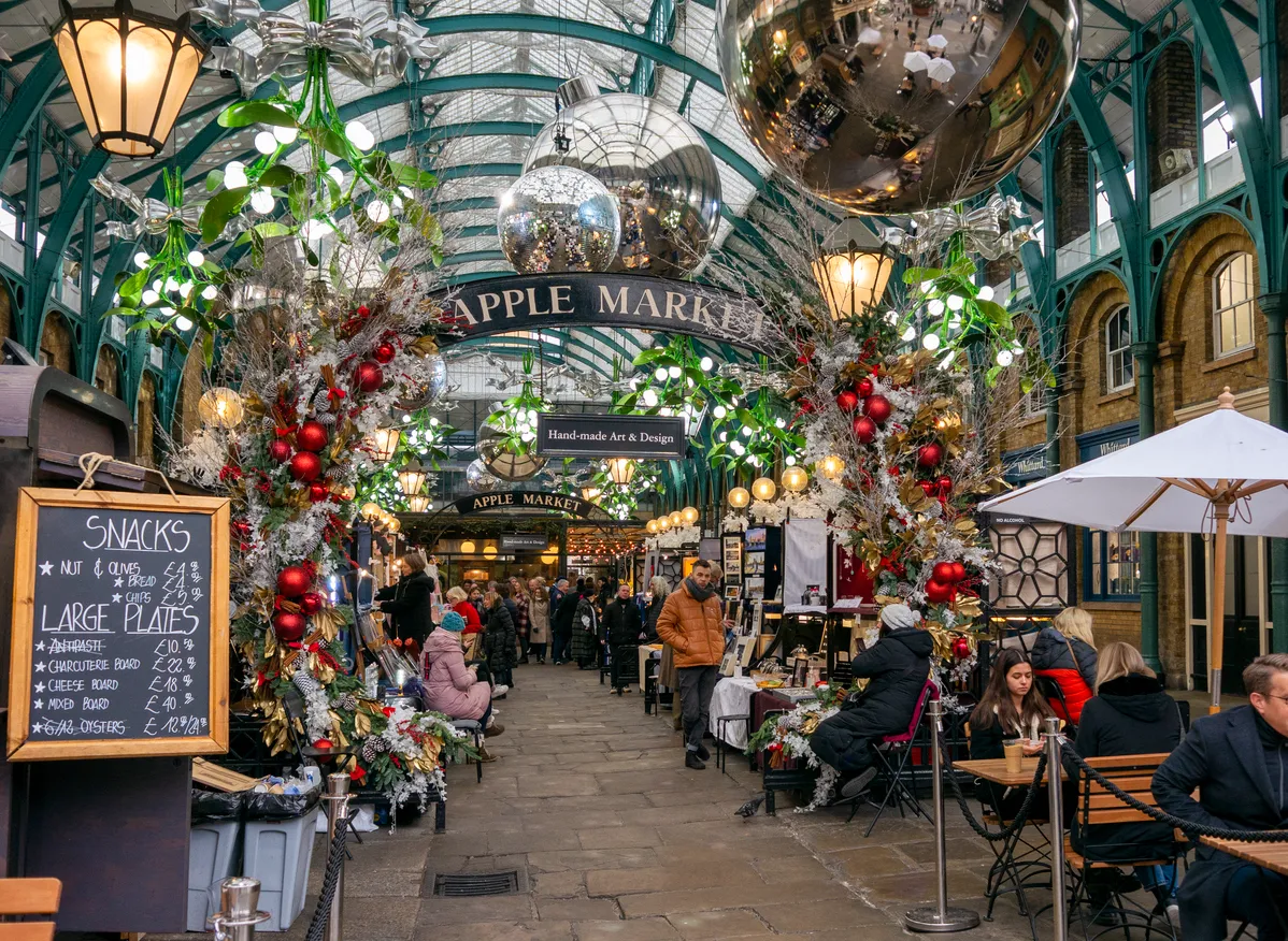 Covent Garden'daki Apple Market'ın Noel süslemeleriyle ışıl ışıl parladığı, büyük yansıtıcı toplar, kırmızı ve altın rengi çelenklerle dolu kapalı çarşıda kalabalıkların alışveriş yaptığı ve kafelerde oturduğu hareketli bir kış sahnesi görülüyor.