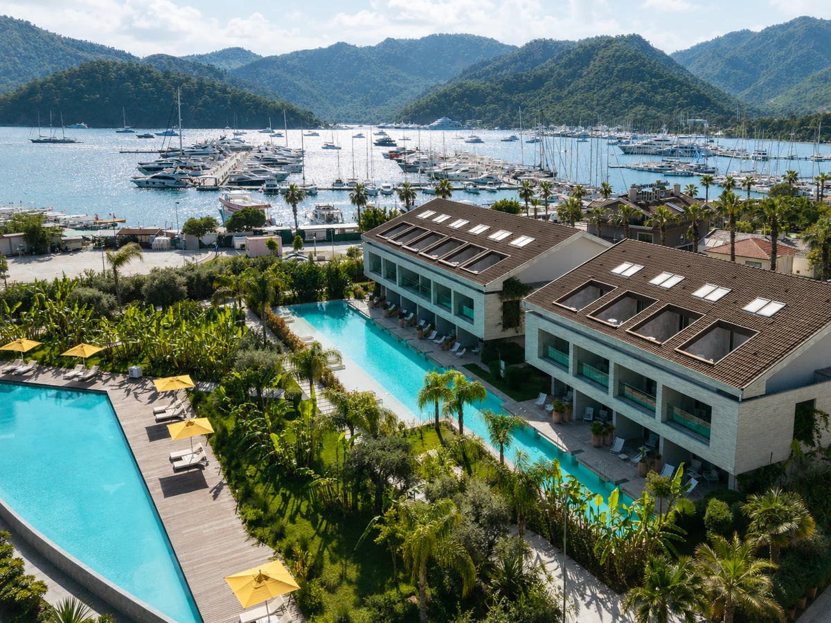 Yat turu için uygun olan Göcek'teki D-Resort Otel'in havadan çekilmiş bu büyüleyici görüntüsünde, yemyeşil tropikal bitki örtüsü ve ahşap zeminlerle çevrili uzun, masmavi yüzme havuzları modern otel binalarının önünde uzanırken, arka planda sayısız lüks yatın demirlediği berrak bir marina ve çam ağaçlarıyla kaplı yeşil tepeler görünüyor.
