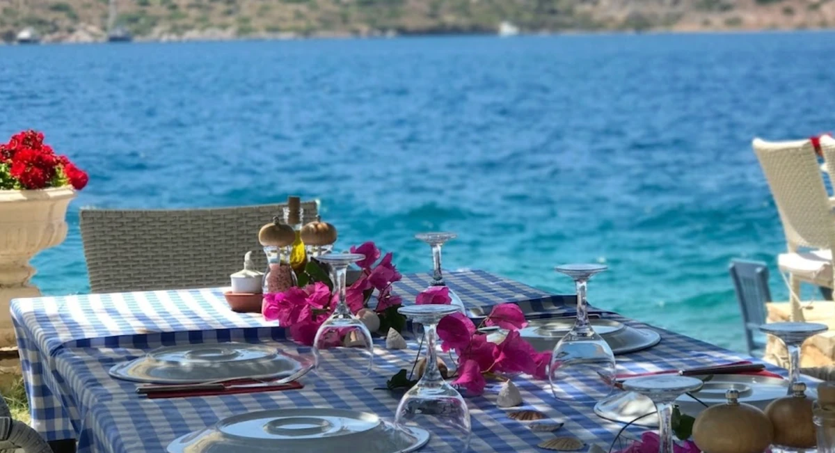 Mavi beyaz kareli bir masa örtüsüyle donatılmış, üzerinde tabaklar, kadehler, pembe begonviller ve deniz kabukları bulunan bir masa, masanın arkasında Yat turu için uygun olan Bozburun'un masmavi denizi ve uzaktaki tepeler, huzurlu bir deniz kenarı restoran atmosferi sunuyor.