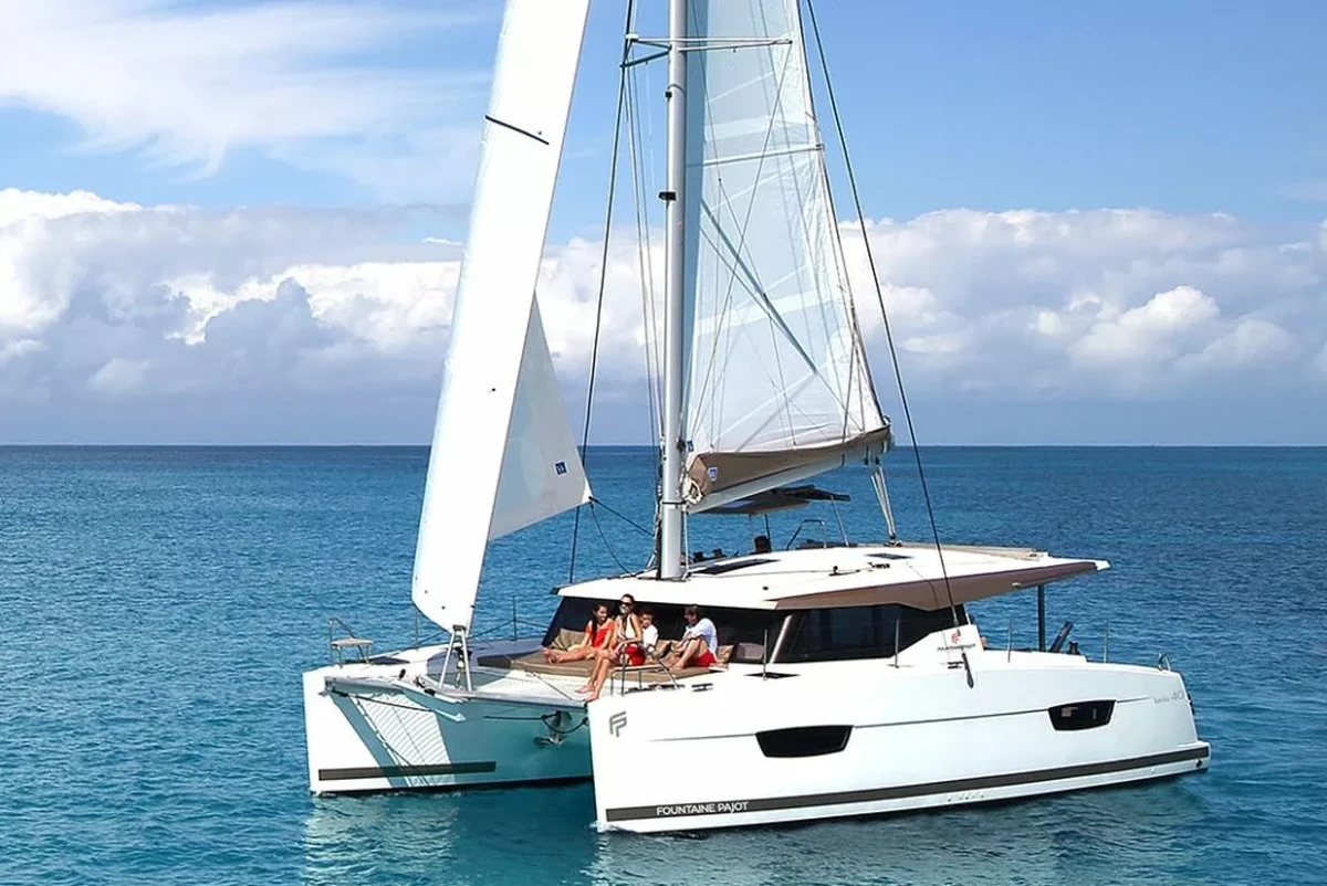 Mavi gökyüzü ve hafif bulutlar altında, berrak turkuaz renkteki denizde ilerleyen Fountaine Pajot marka modern ve şık bir katamaranın ön güvertesinde güneşlenen dört kişi, huzurlu bir deniz tatilinin keyfini çıkarıyor.