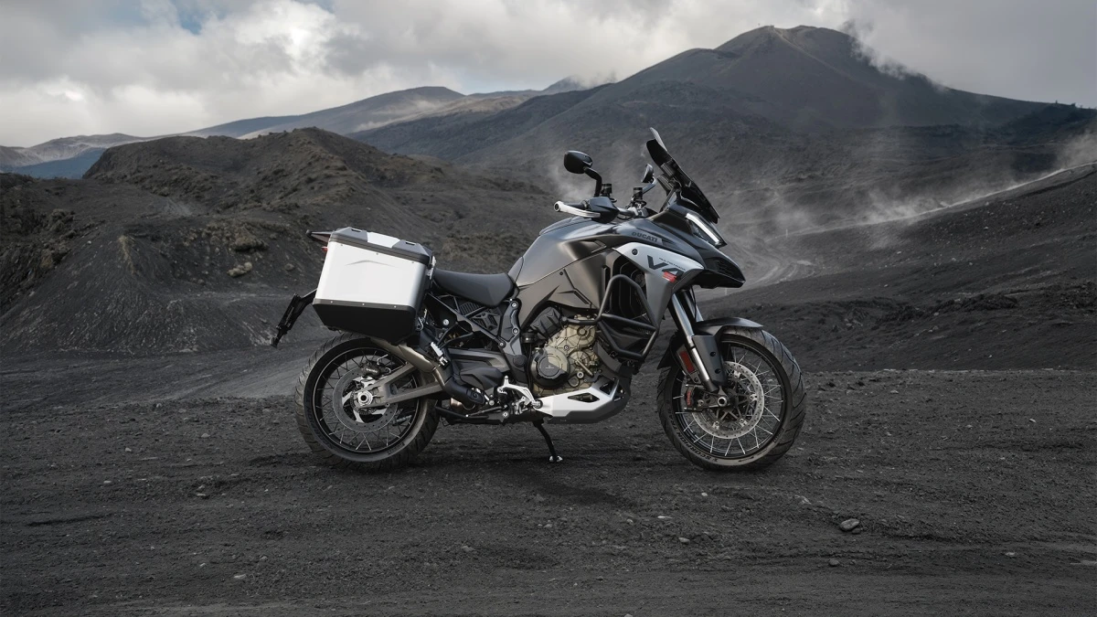 Karanlık, engebeli volkanik bir arazide, bulutlu gökyüzü ve dumanlı dağlar eşliğinde konumlanmış, sol tarafında beyaz bir gezi çantası olan, siyah renkli güçlü bir Ducati Multistrada V4 S gezi motosikleti, maceraya hazır bir duruş sergiliyor.