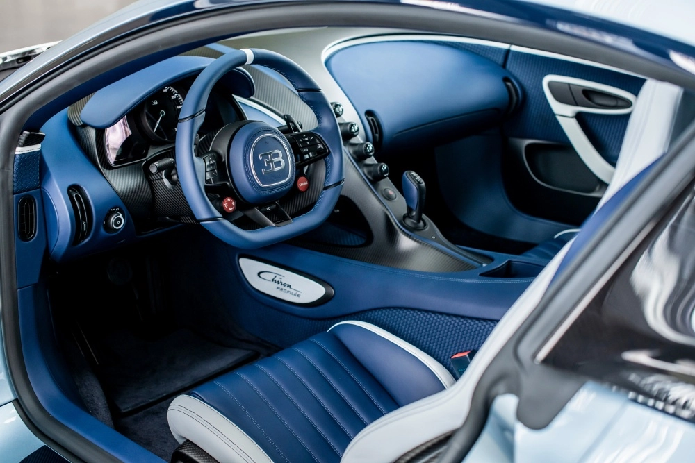 Dünyanın en pahalı arabaları arasında öne çıkan Bugatti Chiron Profilée hiper otomobilin iç mekanından bir görünüm; mavi deri döşemeli spor direksiyonu, karbon fiber detaylarla zenginleştirilmiş gösterge paneli ve orta konsolu ile lüks ve performansı bir arada sunan sofistike tasarımı sergileniyor.