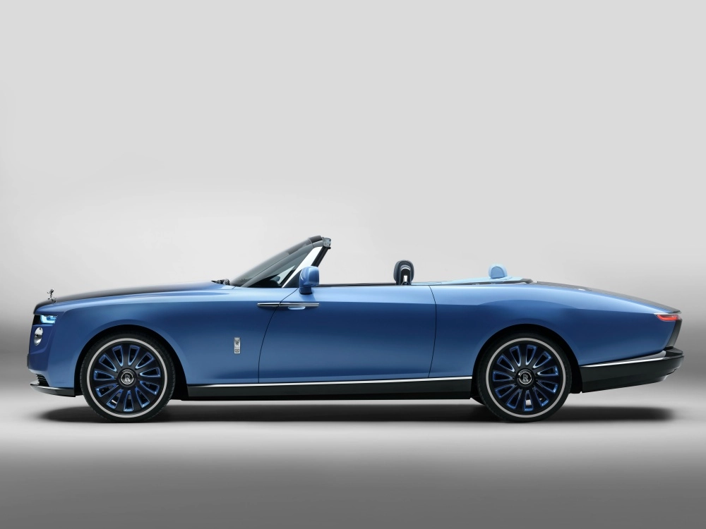 Açık gri bir stüdyo zemininde sol profilden görülen, parlak mavi rengi ve tavanı açık şık tasarımıyla dikkat çeken Rolls-Royce Boat Tail cabrio otomobil, dünyanın en pahalı arabaları arasında öne çıkan el işçiliği ve lüks detaylarını sergiliyor.