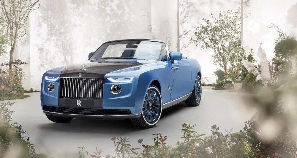 Lüks ve el işçiliğiyle öne çıkan, dünyanın en pahalı arabalarından Rolls-Royce Boat Tail mavi cabrio, siyah kaputu ve parlak ızgarasıyla mistik bir bahçede, yeşil bitkiler ve beyaz tavus kuşu heykelleri arasında tüm ihtişamıyla duruyor.