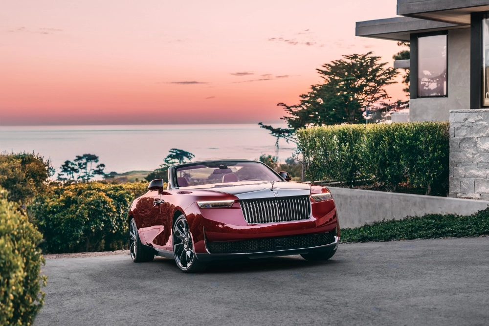 Koyun kırmızı renkteki Rolls-Royce La Rose Noire Drop Tail, gün batımının kızıla boyadığı okyanus manzaralı lüks bir konutun önünde, benzersiz dış tasarımını zarifçe sergileyerek park halinde duruyor.