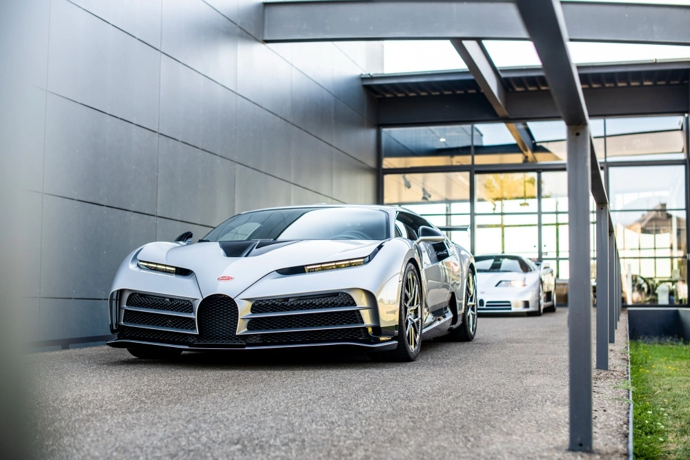 Gümüş renkli, fütüristik tasarımlı Bugatti Centodieci hiper otomobil, modern bir binanın önündeki asfalt zeminde park edilmiş, arkasında da hafif bulanık bir Bugatti EB110 S modeli seçiliyor ve bu özel araçlar dünyanın en pahalı otomobillerinden ikisi olarak öne çıkıyor.