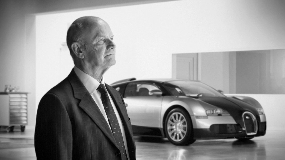 Bugatti Veyron'un arkasındaki mühendislerden Ferdinand Karl Piëch'in siyah beyaz fotoğrafında, takım elbiseli Piëch dikkatle sağa bakarken, iki tonlu lüks spor araba Veyron bulanık bir şekilde arka planda sergileniyor.