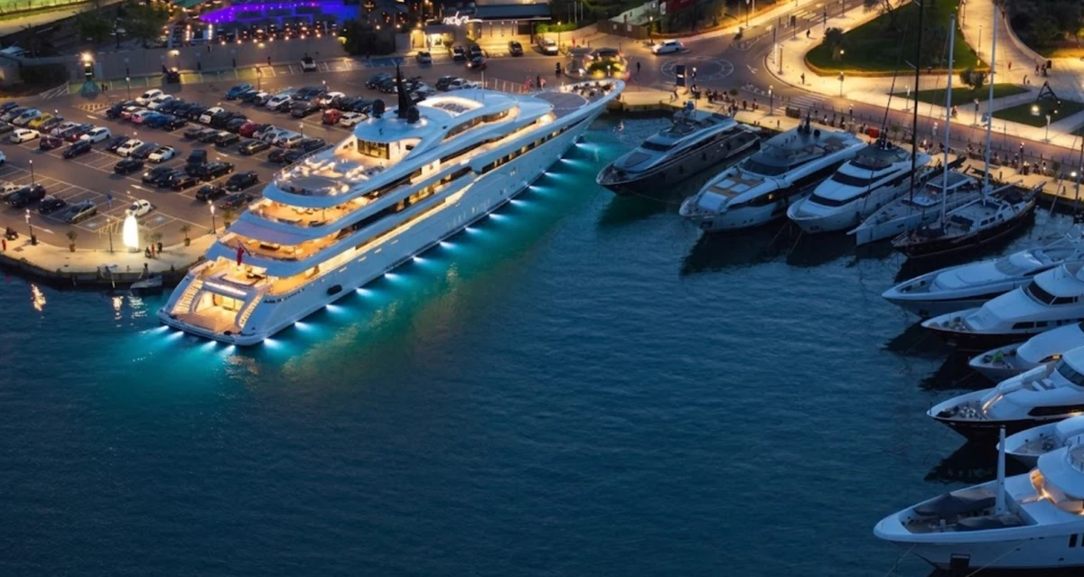 Yat turu için uygun olan D-Marin Turgutreis'in hareketli gece marinasında, suya yansıyan mavi alt ışıklarıyla büyüleyici bir süperyatın yanı sıra lüks yatlar ve yelkenliler demirlemiş, karadaki aydınlatılmış otopark ve binalar da manzarayı tamamlıyor.