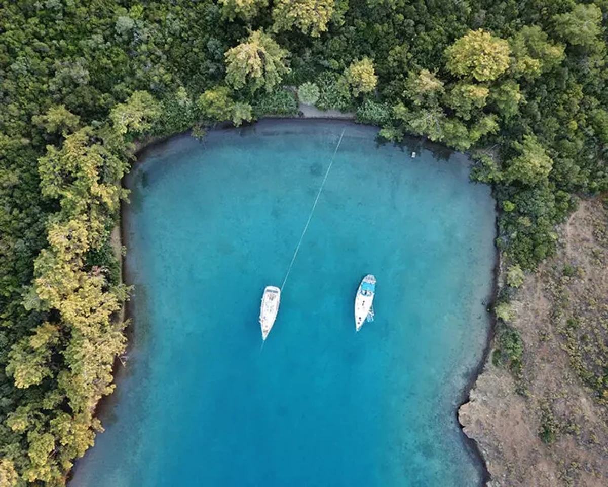 Göcek'in yemyeşil ağaçlarla çevrili kıyı şeridini ve turkuaz sularında sakin sakin sallanan iki beyaz yelkenli tekneyi gösteren bu havadan görünüm, bölgenin dingin ve doğal güzelliğini vurguluyor.