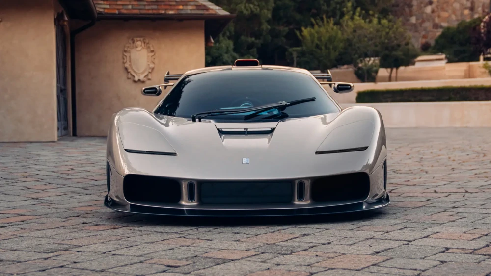 Ön planda, Gordon Murray S1 LM adlı, şampanya rengi parlak bir hiper otomobilin agresif dış tasarımı önden gösteriliyor; aracın yere yakın geniş gövdesi, dikkat çekici aerodinamik hatları, büyük ön hava girişleri ve tavanındaki turuncu hava kepçesi taş döşeli yolda belirginleşirken, arka planda hafif bulanık bir malikane uzanıyor.