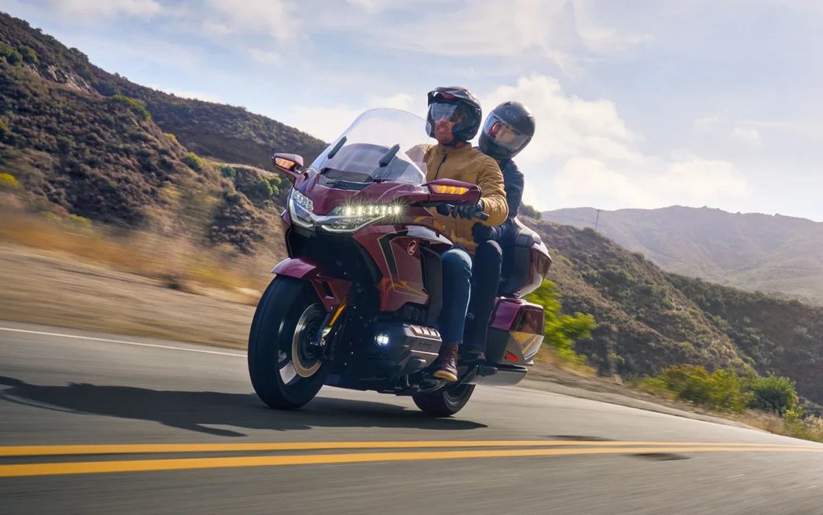 Virajlı dağ yolunda ilerleyen, koyu kırmızı renkteki bir Honda Gold Wing gezi motosikleti üzerinde, kask takmış bir sürücü ve yolcusu, parlak gökyüzü altında keyifli bir sürüş yaparken görülüyor, uzun yolculuklara uygun tasarımıyla dikkat çekiyor.