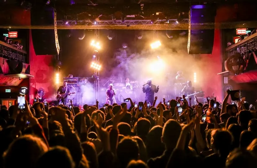 Jolly Joker Ankara mekanında sahne ışıkları ve duman efektleri altında canlı müzik performansı sergileyen bir müzik grubu ve konseri telefonlarıyla kaydeden elleri havada coşkulu bir kalabalık görülüyor.